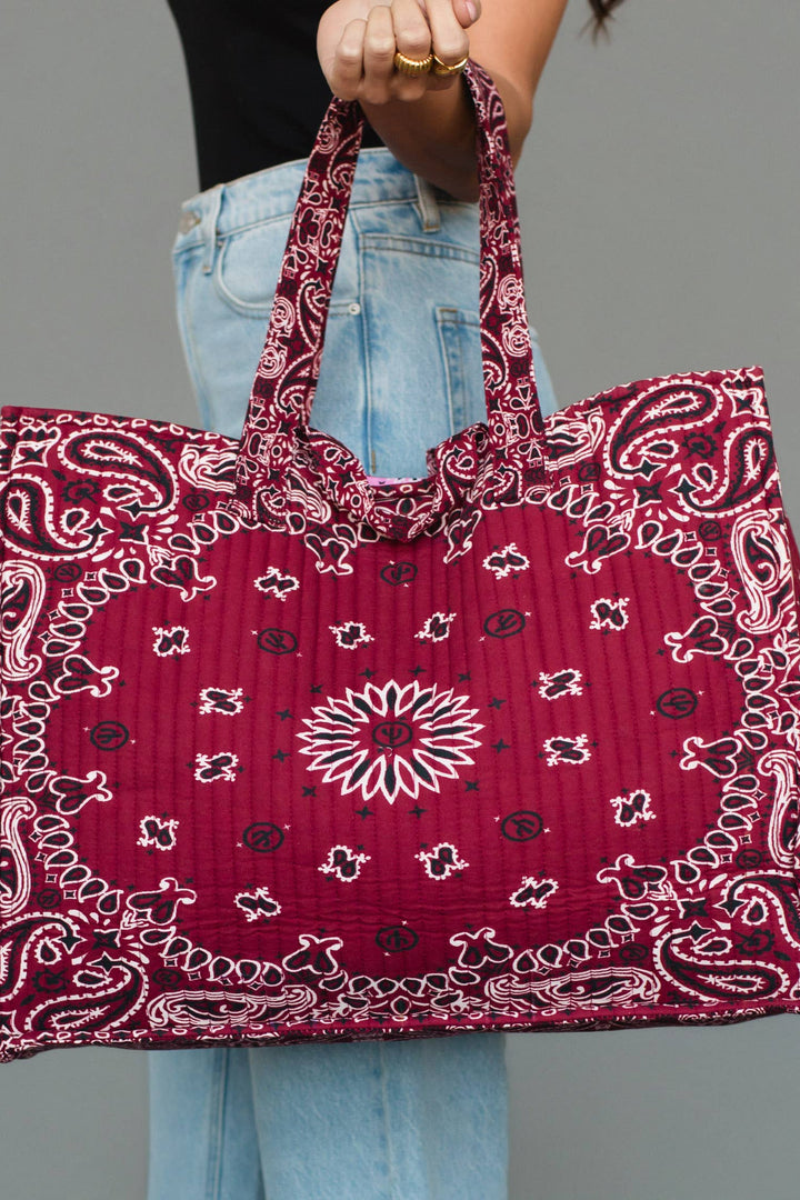 A person holding a large quilted tote bag with a red paisley bandana pattern, shown against a plain gray studio background.