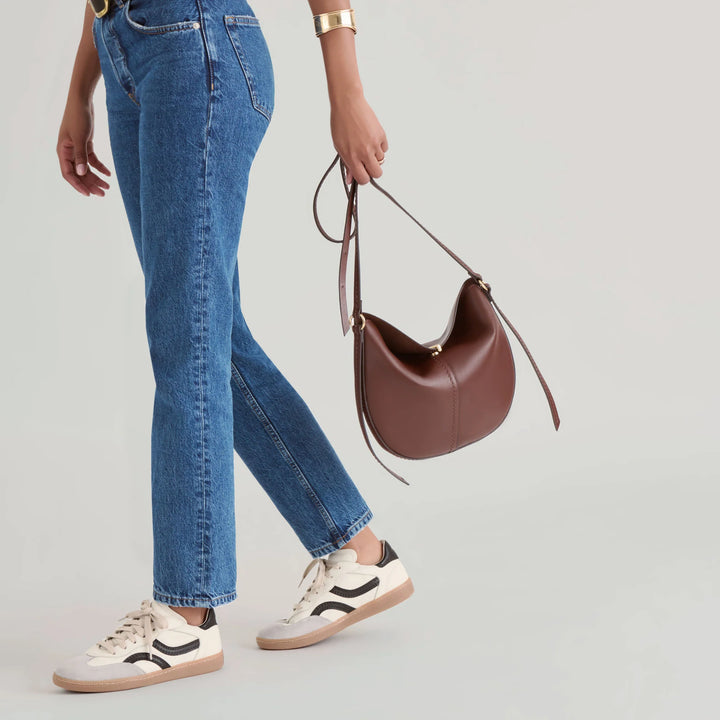 Person wearing blue jeans and white sneakers holding a brown leather handbag 