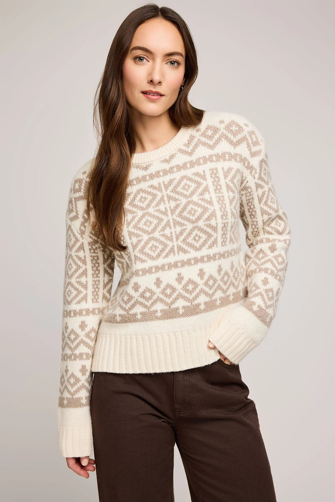 Woman wearing a cream patterned sweater and dark pants against a plain background