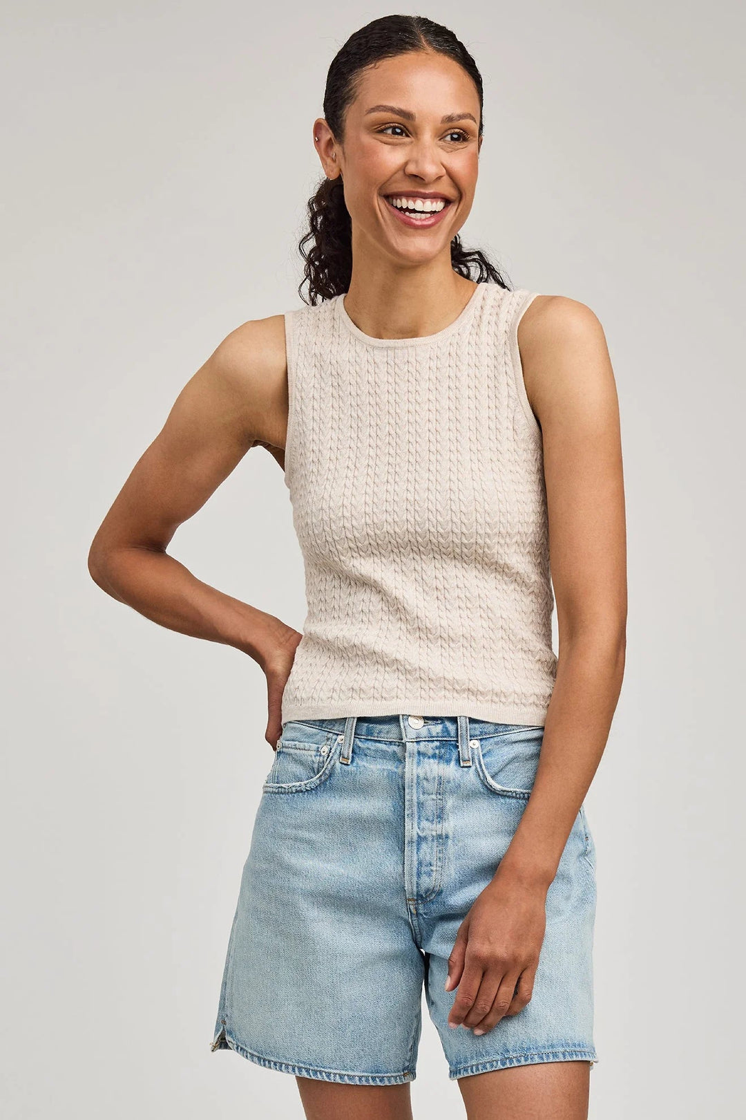 Woman wearing a beige sleeveless top and light blue denim shorts on a plain background