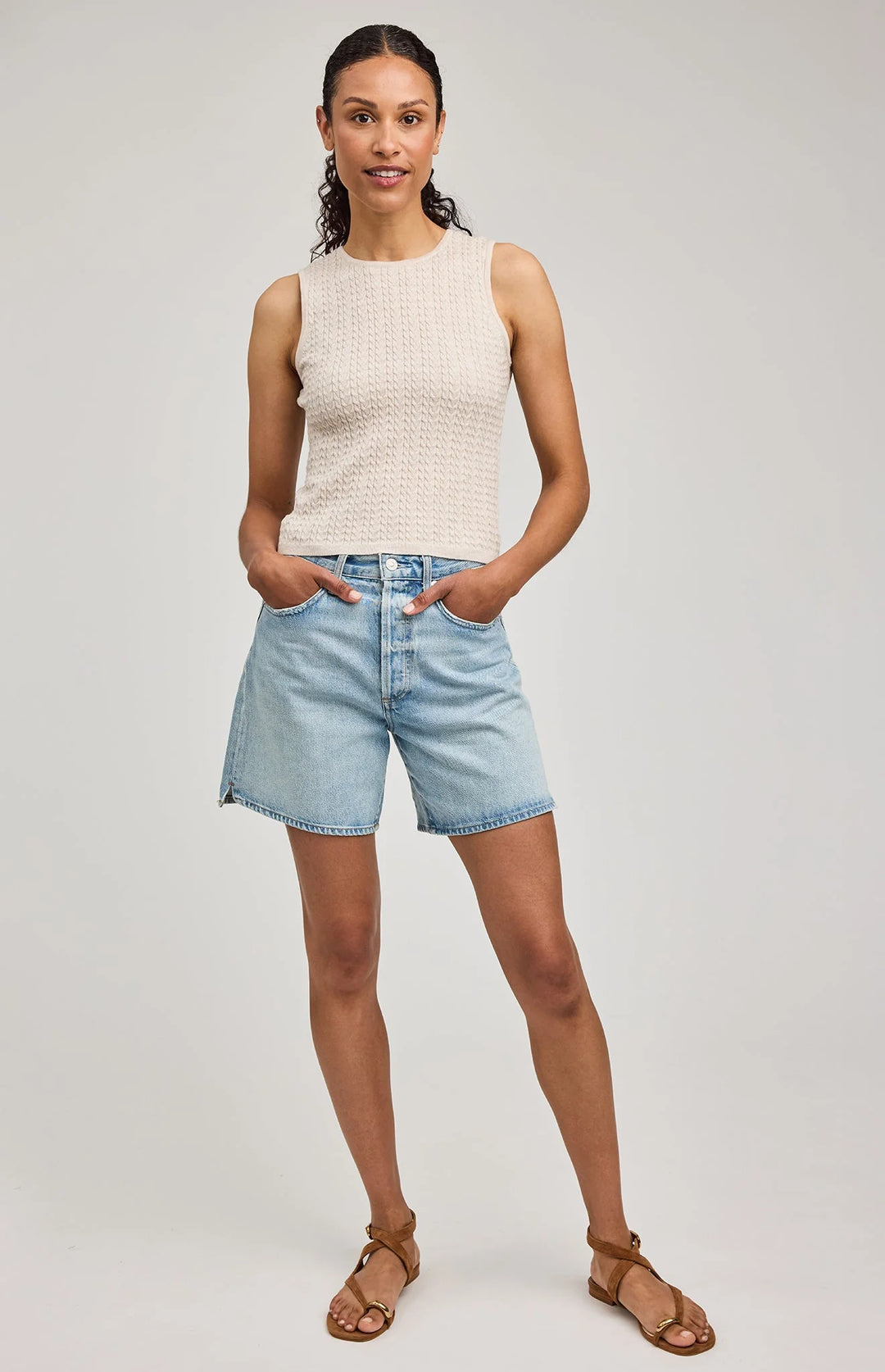 Woman wearing a sleeveless beige top and light blue denim shorts on a plain background
