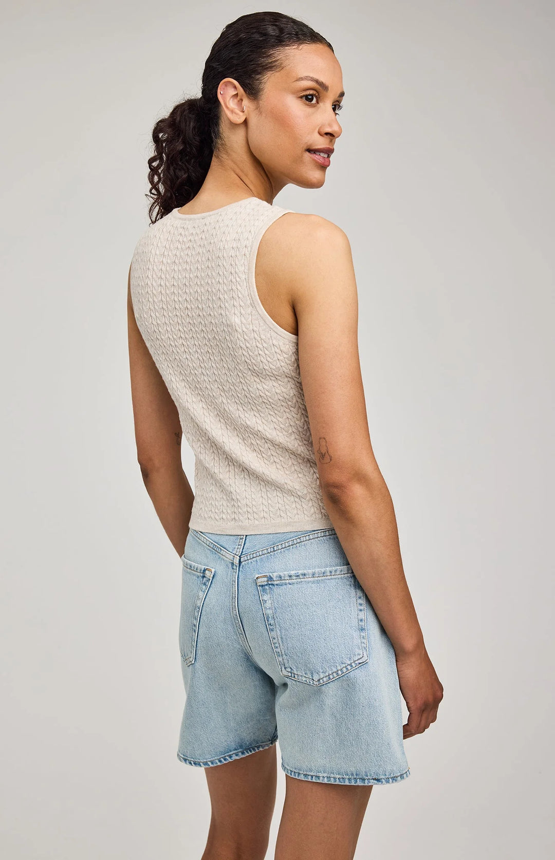 Woman wearing a beige sleeveless top and light blue denim shorts on a plain background