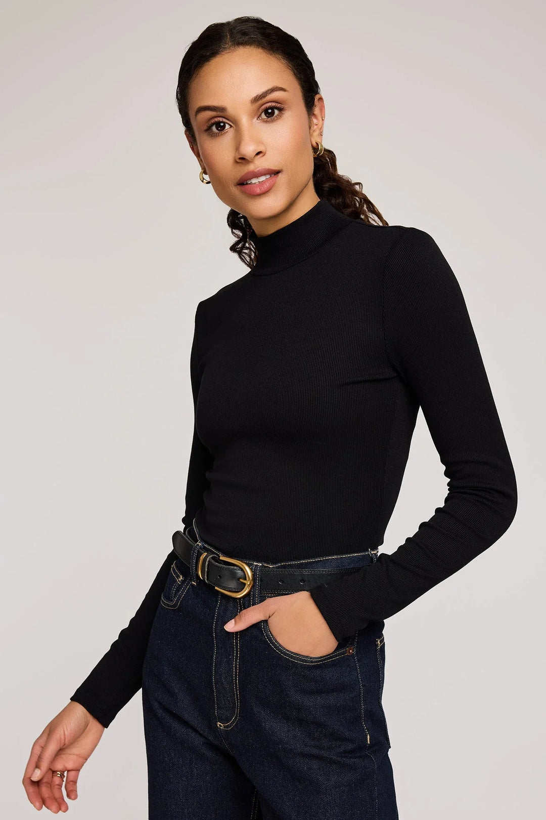 Woman wearing a black turtleneck and dark jeans against a plain background