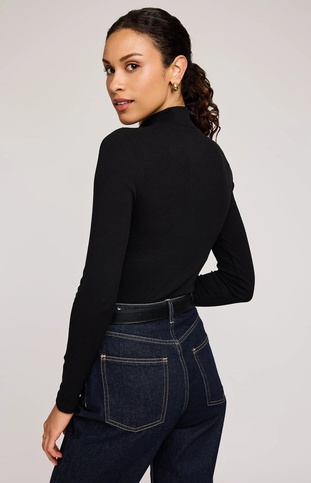 Woman wearing a black long-sleeve top and dark jeans on a plain background