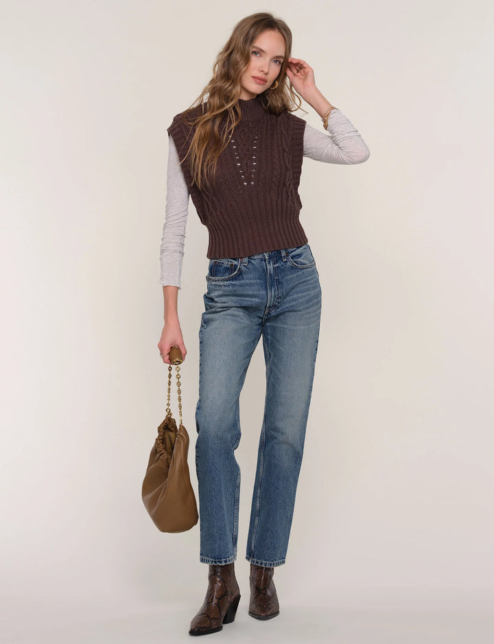 Woman wearing a brown knitted vest, light gray long-sleeve shirt, blue jeans, and brown boots, holding a brown handbag on a white background.