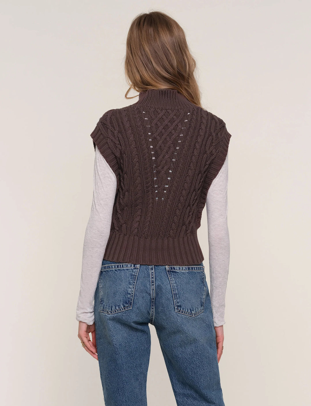 Person wearing a brown knitted vest over a light gray long-sleeve shirt and blue jeans on a plain background