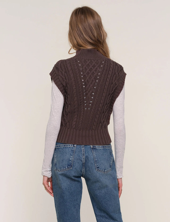 Person wearing a brown knitted vest over a light gray long-sleeve shirt and blue jeans on a plain background