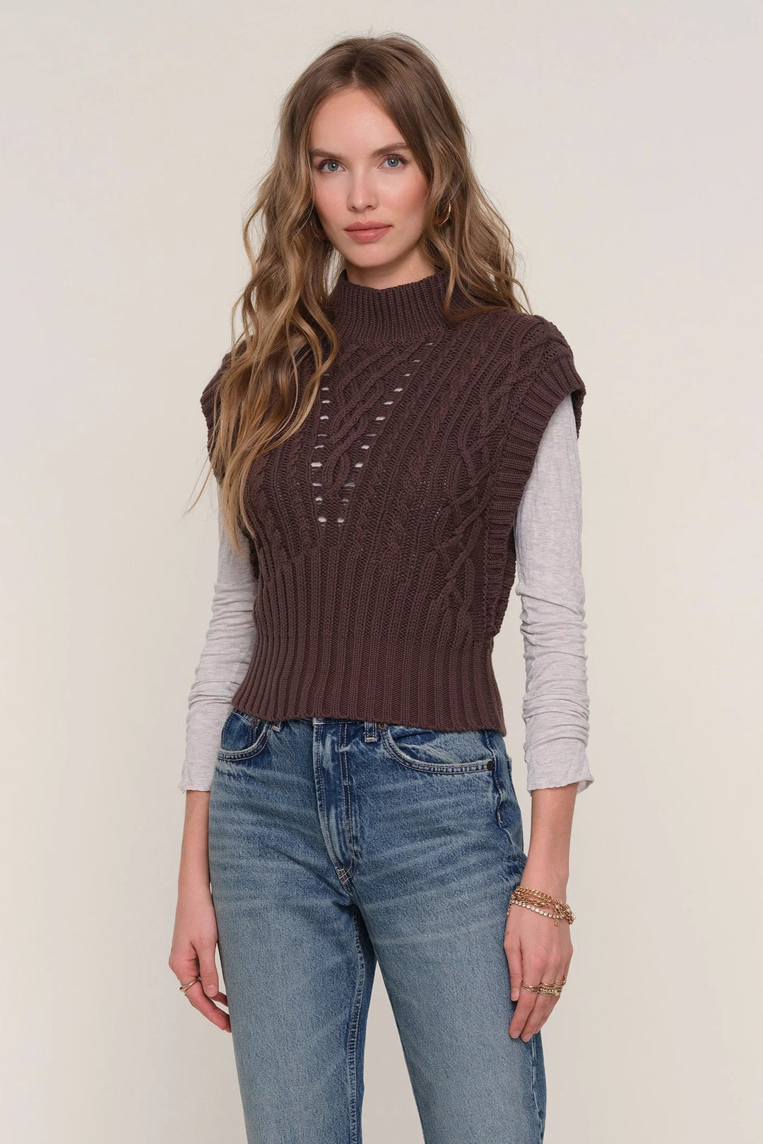 Woman wearing a brown knitted vest over a light gray shirt with blue jeans on a plain background