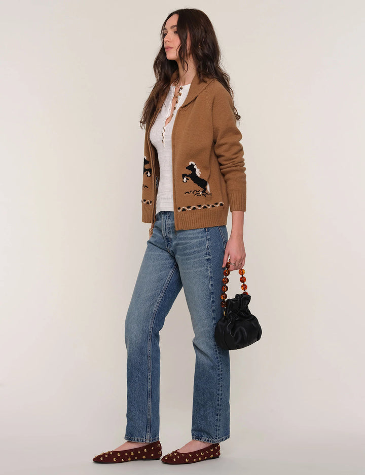 Woman wearing a brown cardigan with a pattern, white shirt, blue jeans, and brown shoes on a plain background.