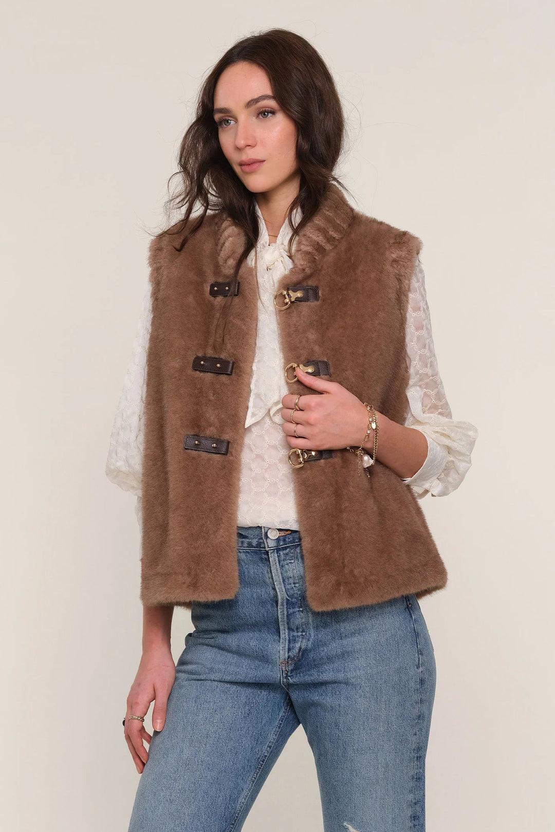 Woman wearing a brown vest over a white shirt and blue jeans on a plain background