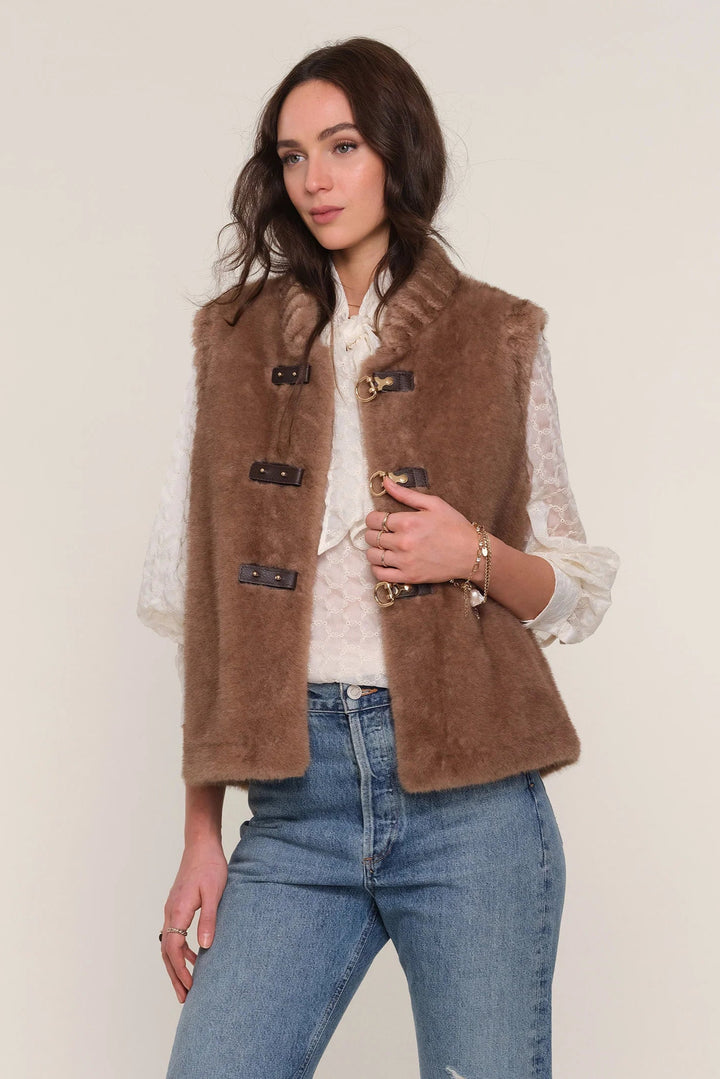 Woman wearing a brown vest over a white shirt and blue jeans on a plain background