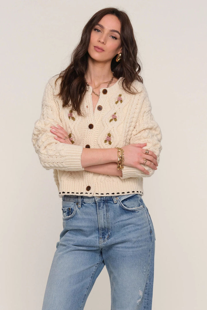 Woman wearing a cream cardigan with black buttons and blue jeans on a plain background