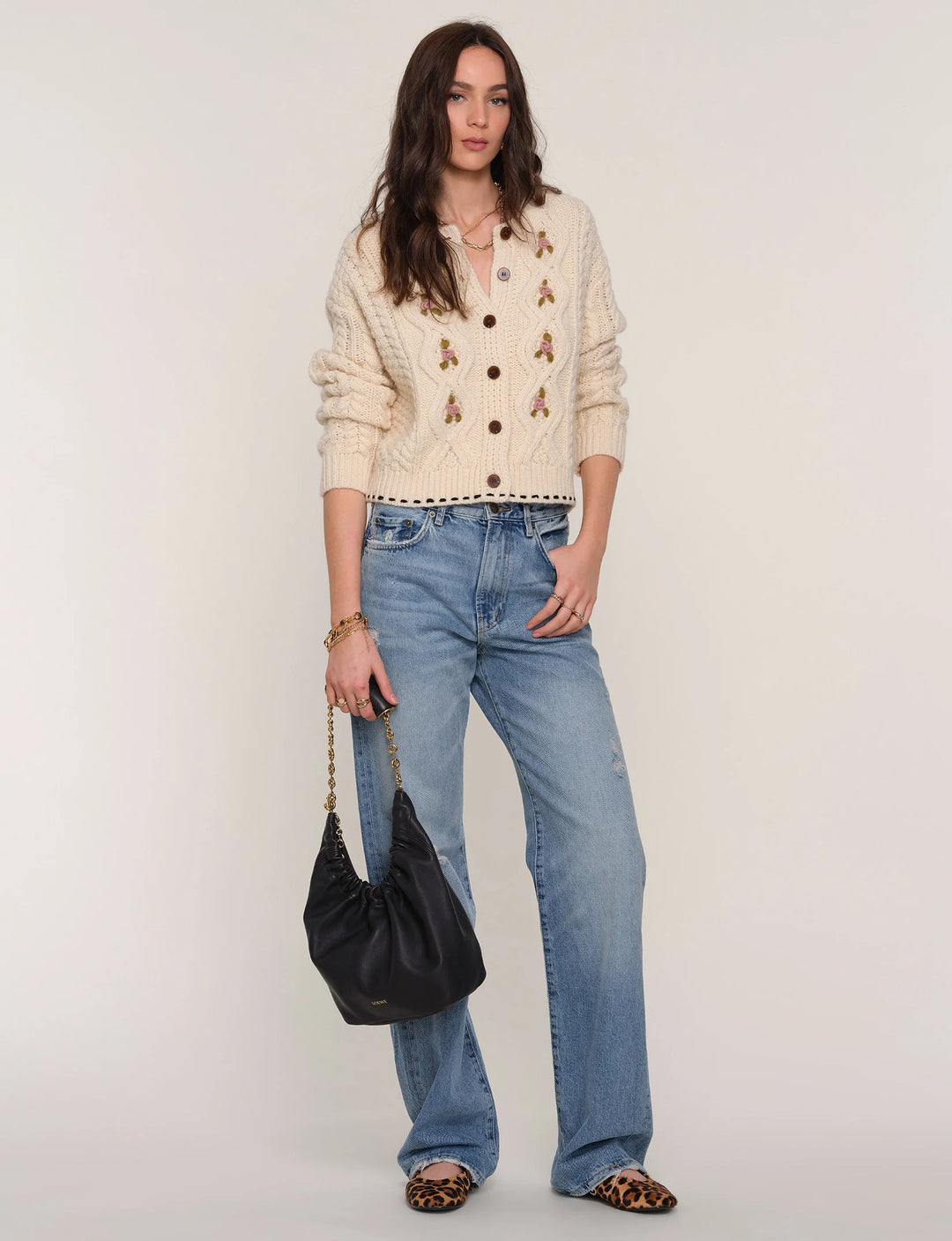 Woman wearing a beige cardigan, blue jeans, and holding a black handbag on a white background