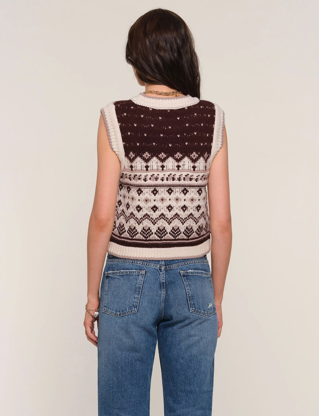 Person wearing a patterned sleeveless sweater and blue jeans on a plain background