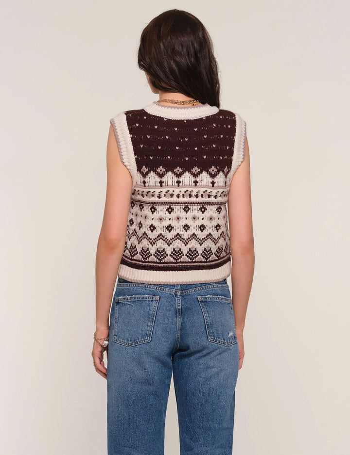 Person wearing a patterned sleeveless sweater and blue jeans on a plain background