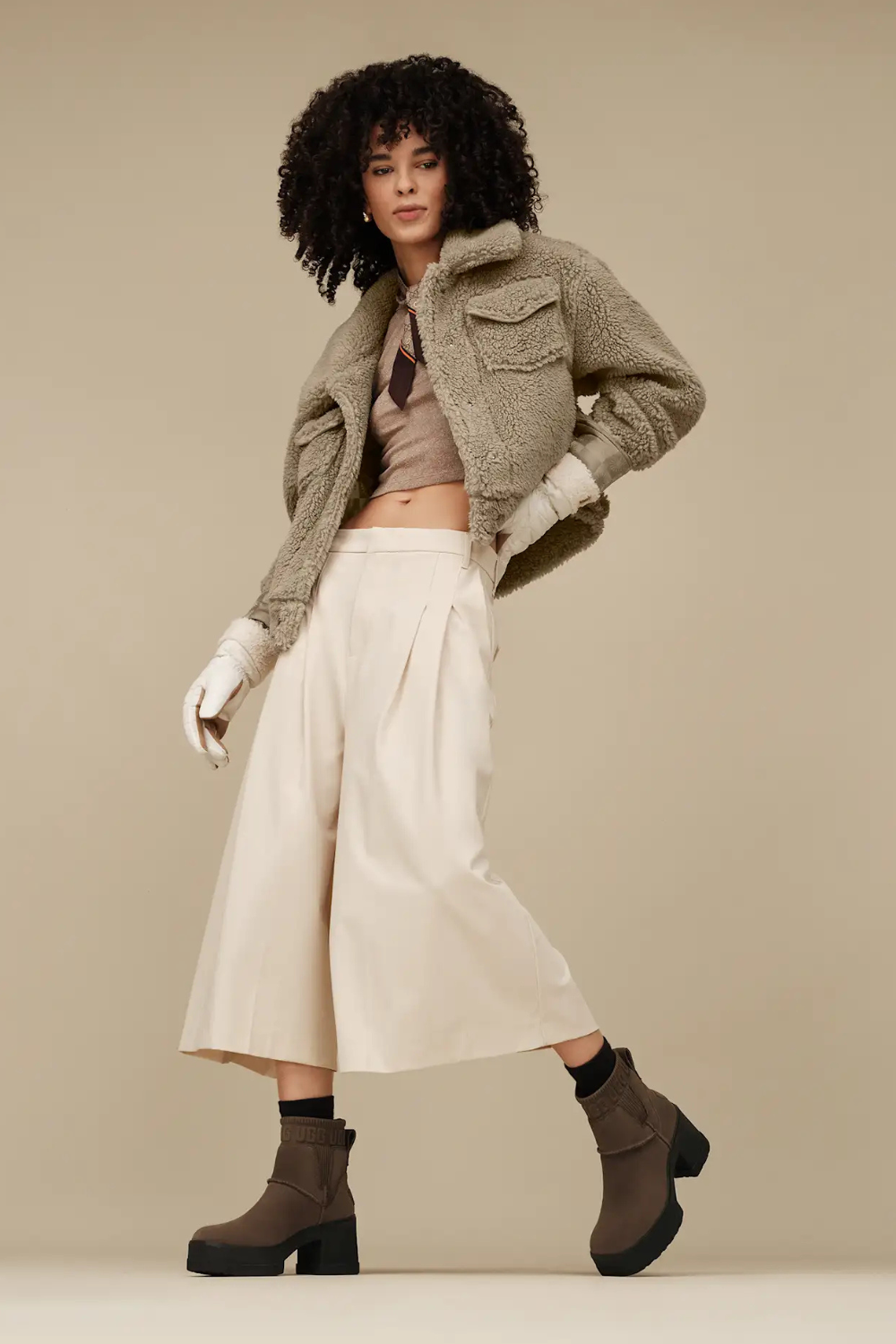 Woman wearing a green jacket, white pants, and brown suede ugg Chelsea boots on a beige background