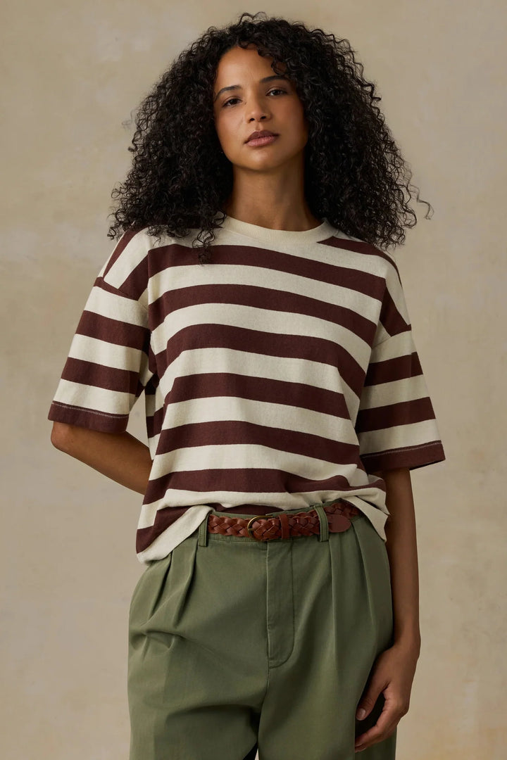 A woman with curly dark hair wearing a brown and cream horizontally striped short-sleeve t-shirt, olive green trousers, and a brown woven belt, standing against a textured beige background.