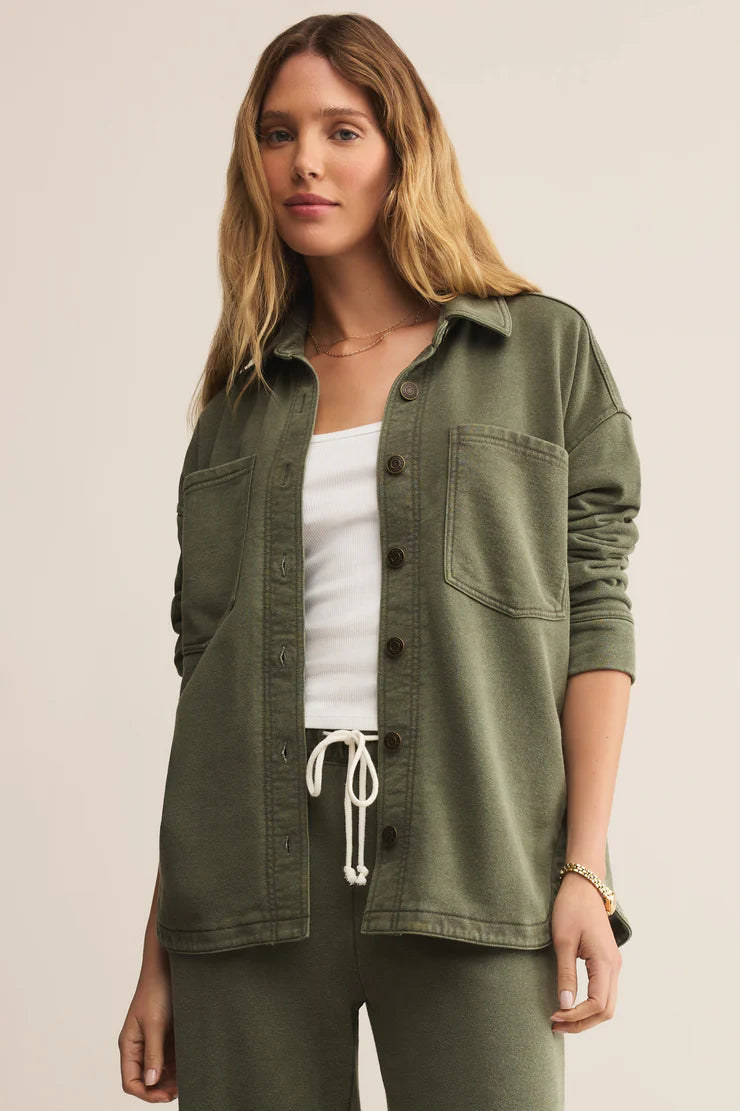 Woman wearing an olive green button-up shirt over a white top and green pants on a beige background