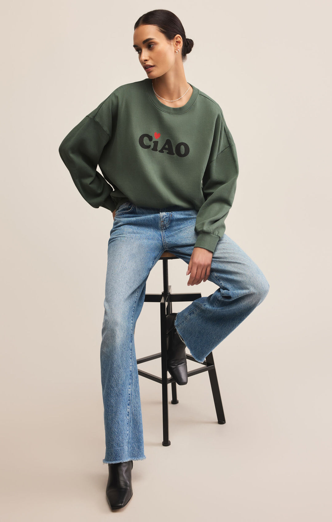 Woman wearing a green sweatshirt with 'CIAO' text, sitting on a stool against a beige background
