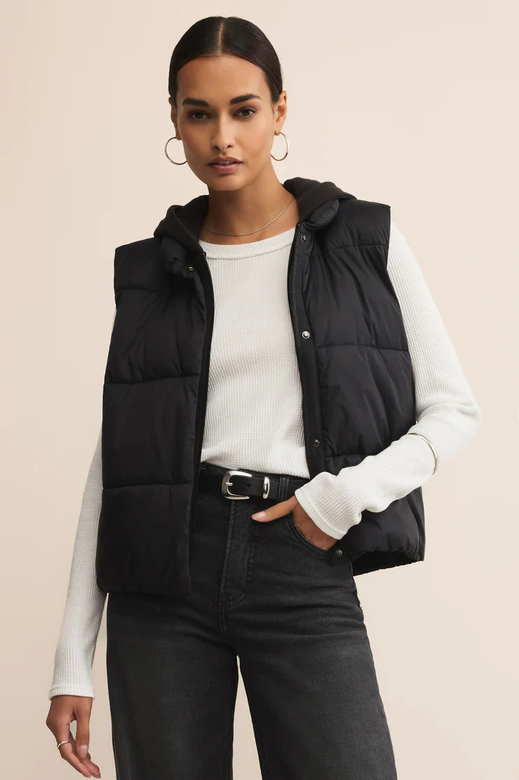 Woman wearing a black puffer vest over a white long-sleeve shirt on a beige background
