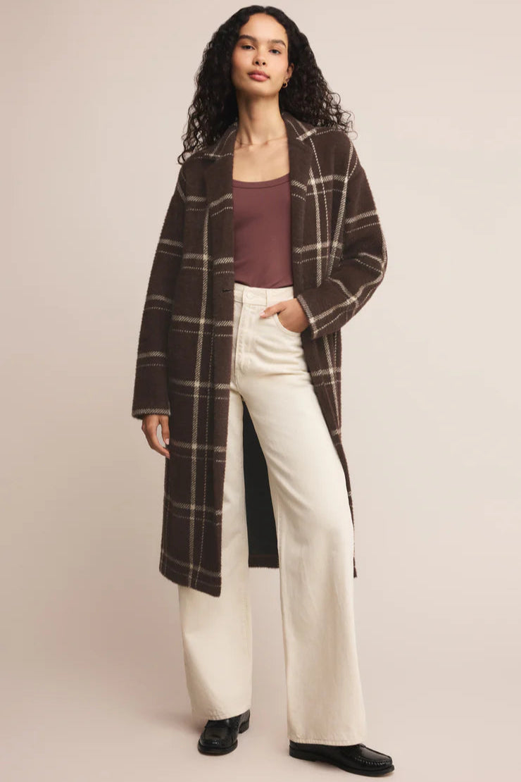 Woman wearing a plaid coat, purple top, and white pants on a beige background
