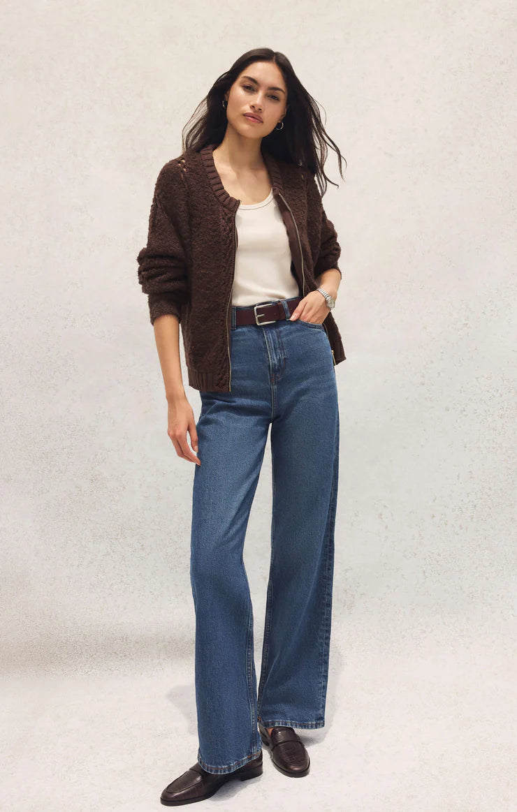 Woman wearing a brown cardigan, white shirt, blue jeans, and black shoes on a light gray background