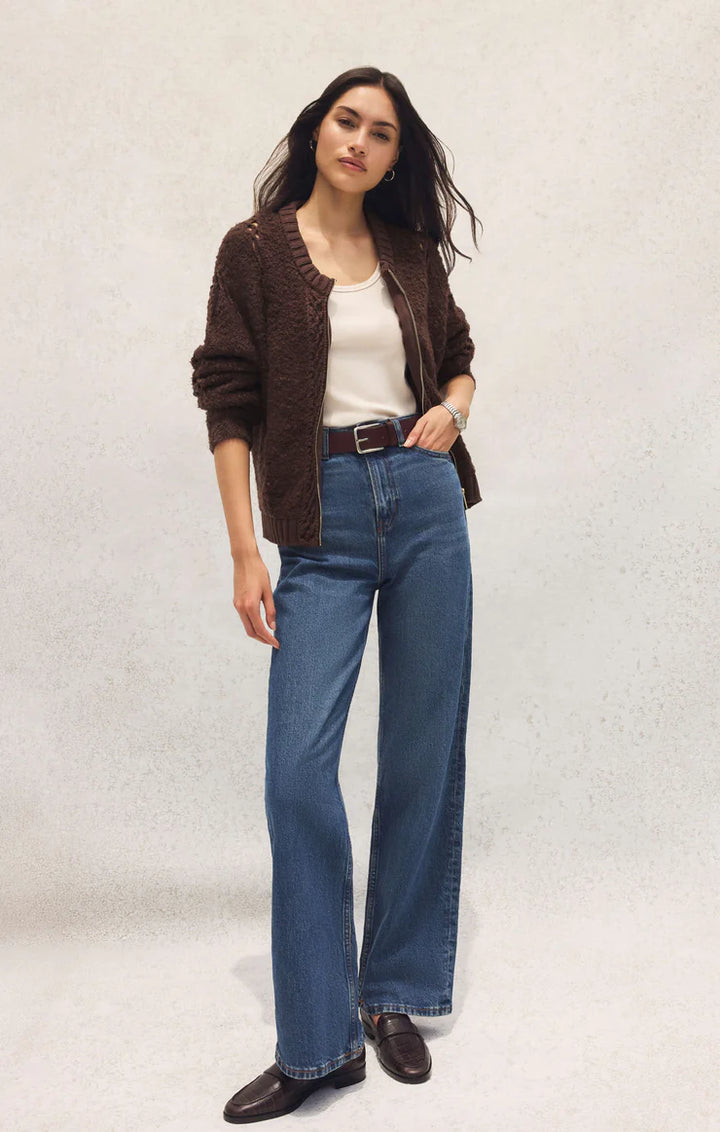 Woman wearing a brown cardigan, white shirt, blue jeans, and black shoes on a light gray background